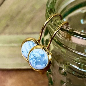 May include: A pair of gold-coloured earrings featuring round, light blue and white marbled stone centres. The earrings have a simple, elegant design with a lever back closure. The earrings are displayed against a blurred green background.