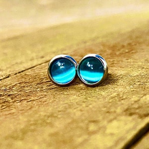 May include: A pair of silver stud earrings with blue glass cabochons. The cabochons have a gradient of blue, fading from a darker blue to a lighter blue.