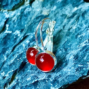 May include: A pair of silver-toned leverback earrings with round, vibrant red cabochon stones. The earrings are set against a textured, dark blue-gray surface, highlighting the polished metal and the rich color of the stones.