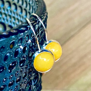 May include: A pair of silver-tone earrings with round, yellow-colored stones. The earrings have a simple hook design and are set against a blurred background. The earrings are a jewelry item.
