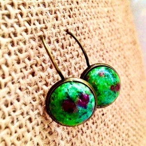 May include: A pair of round earrings featuring a green and red speckled design. The earrings have a bronze-coloured metal frame and a curved hook. The background is a tan, textured fabric.