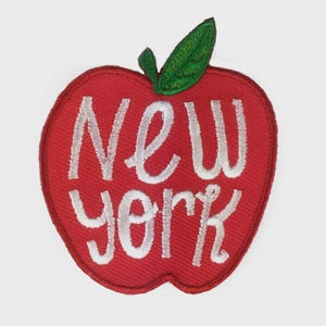 Pode incluir: Um patch vermelho em forma de maçã com as palavras "NEW YORK" bordadas em branco. O patch tem uma folha verde no topo. O patch é provavelmente para roupas ou acessórios.