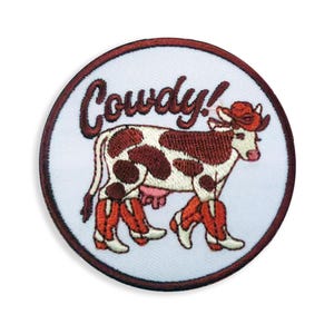 Könnte beinhalten: Gestickter Aufnäher mit einer Kuh, die einen Cowboyhut und Stiefel trägt. Die Kuh hat braune Flecken und befindet sich vor einem weißen Hintergrund mit dem Wort "Cowdy!" in brauner Schrift. Der Aufnäher hat einen braunen Rand.