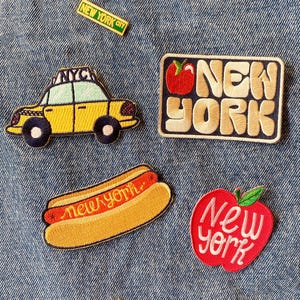 May include: A collection of five embroidered patches featuring a yellow taxi with NYC text, a rectangular patch with the text NEW YORK and a red apple, a hot dog with new york text, a red apple with New York text, and a small NEW YORK CITY pin.