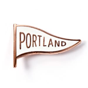 May include: Rose gold-colored metal pin shaped like a pennant with a white background and the word "PORTLAND" in black lettering.