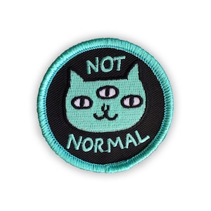 May include: A round, black and teal embroidered patch with a green cat with three eyes and the text "NOT NORMAL" in white.