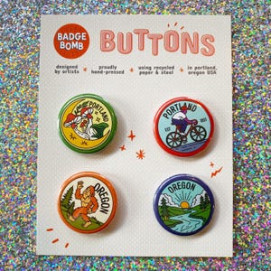 May include: A set of four circular pin-back buttons on a white display card labeled Badge Bomb Buttons. The buttons feature illustrations of mushrooms, a person on a bicycle, a sasquatch, and a landscape with a river, trees, and sun.