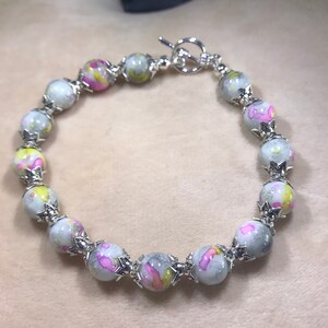 May include: A silver bracelet with a toggle clasp features round, white beads with pink, yellow, and gray marbling. Each bead is accented with a silver star-shaped charm.