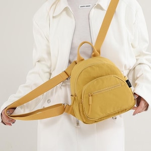 May include: Mustard yellow backpack with a front zippered pocket and a top handle. The backpack has adjustable shoulder straps and a small tag on the side. The bag is made of a lightweight material.