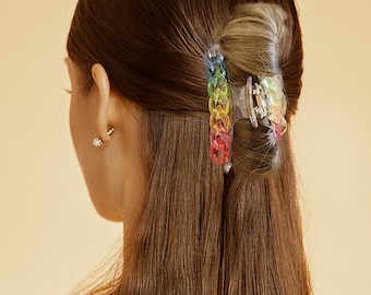 May include: A hair claw clip featuring a translucent base and a decorative outer edge composed of interlocking, multi-colored plastic links in a vibrant rainbow gradient of blue, green, yellow, orange, and pink, securing a section of brown hair.