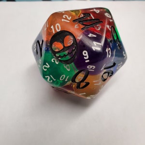 Pode incluir: Dado de 20 faces de plástico transparente multicolor com números pretos e um gráfico em preto e branco de um rosto de monstro. O dado mostra o número 16.