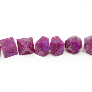 Könnte beinhalten: Satz von sieben pinkfarbenen und violetten, schillernden Würfeln mit Glitzer, der in das Harz eingebettet ist. Die Würfel sind von 1 bis 6 nummeriert.