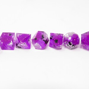 Könnte beinhalten: Satz von sieben lila und schwarzen Würfeln mit Glitzer. Die Würfel sind durchsichtig und haben schwarze Zahlen und weiße Punkte.