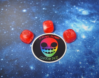 Może przedstawiać: Trzy czerwone kostki do gry z czarnymi kropkami na niebieskim tle z białymi gwiazdami. Naklejka z tęczową twarzą kosmity i tekstem "Normal Human Custom Dice" znajduje się w środku.