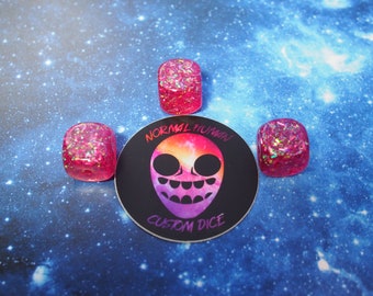May include: Three pink dice with iridescent glitter embedded in the resin. The dice are sitting on a blue background with a sticker that says "Normal Human Custom Dice".