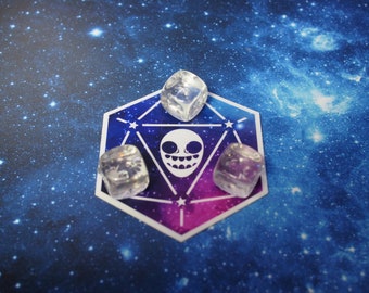 May include: Three clear, cube-shaped dice rest on a hexagonal sticker with a white skull design and a blue, purple, and pink background. The sticker is on a blue background with white stars.