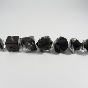 May include: Set of seven black and clear dice with white numbers. The dice are made of a translucent material with a black swirl.