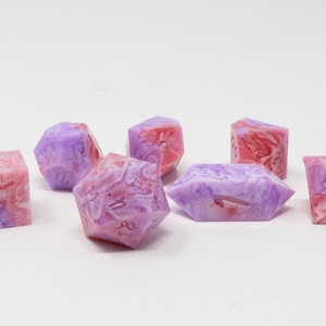 以下が含まれることがあります： 紫、ピンク、白の色合いの大理石模様のサイコロ7個セット。サイコロは、立方体、d20、d6など、さまざまな形をしています。サイコロは半透明の素材でできています。