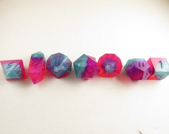 May include: Set of seven iridescent dice with pink and blue glitter embedded in the clear resin. The dice are numbered 1 through 7.