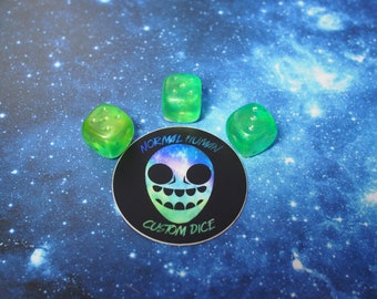 May include: Three green and blue dice with a galaxy design on a black sticker that says "Normal Human Custom Dice".