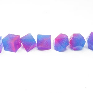 May include: Set of seven iridescent dice with a pink and blue galaxy swirl pattern. The dice are made of resin and have a glittery finish.