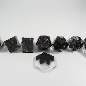 May include: A set of seven black and iridescent dice with white numbers. The dice are arranged in a line on a white surface. The dice are made of resin and have a glittery finish.