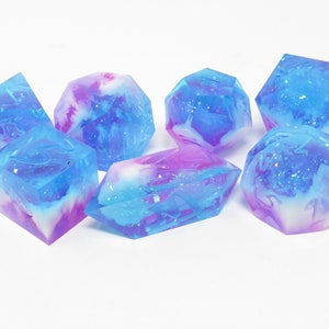Op de afbeelding: Set van zes iriserende dobbelstenen met een blauw en wit swirl-patroon en glitter. De dobbelstenen zijn gemaakt van hars en hebben verschillende vormen, waaronder een kubus, een d4, een d6, een d8, een d10 en een d12.