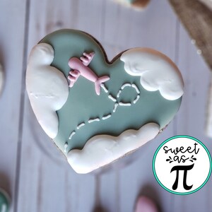 May include: A heart-shaped cookie decorated with green icing, white clouds, and a pink airplane flying through the clouds. The cookie has a dotted line trail behind the airplane. The cookie is on a white wooden surface. The text "sweet as &pi;" is on a blue circle in the lower right corner of the image.