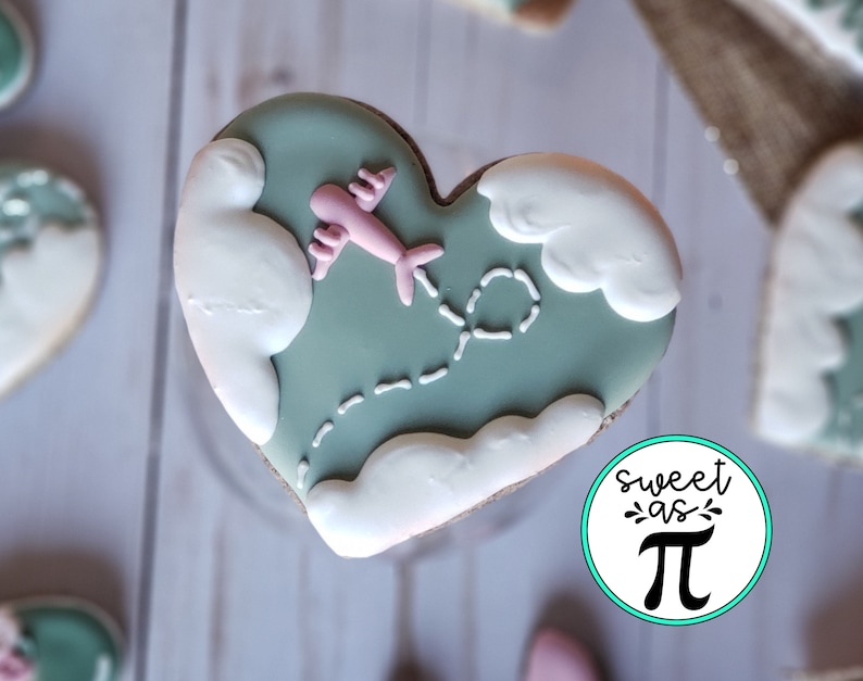 May include: A heart-shaped cookie decorated with green icing, white clouds, and a pink airplane flying through the clouds. The cookie has a dotted line trail behind the airplane. The cookie is on a white wooden surface. The text "sweet as &pi;" is on a blue circle in the lower right corner of the image.