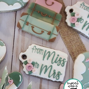 May include: A set of three decorated sugar cookies. The cookies are shaped like suitcases and have a green, pink, and brown color scheme. The cookies are decorated with white icing and have a "from Miss to Mrs" tag attached to one of the cookies.
