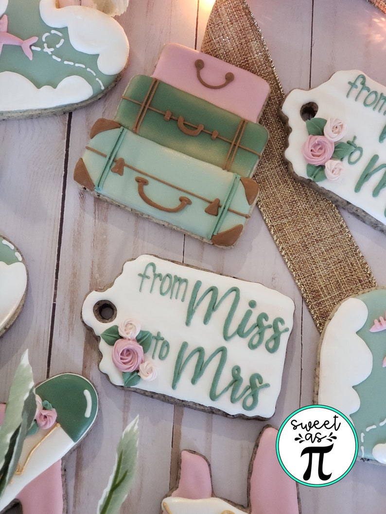 May include: A set of three decorated sugar cookies. The cookies are shaped like suitcases and have a green, pink, and brown color scheme. The cookies are decorated with white icing and have a "from Miss to Mrs" tag attached to one of the cookies.
