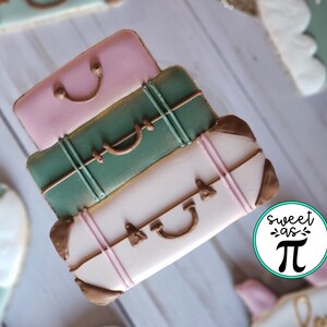 May include: A stack of three decorated cookies shaped like suitcases. The top suitcase is pink, the middle suitcase is green, and the bottom suitcase is white. Each suitcase has a brown handle and gold accents.