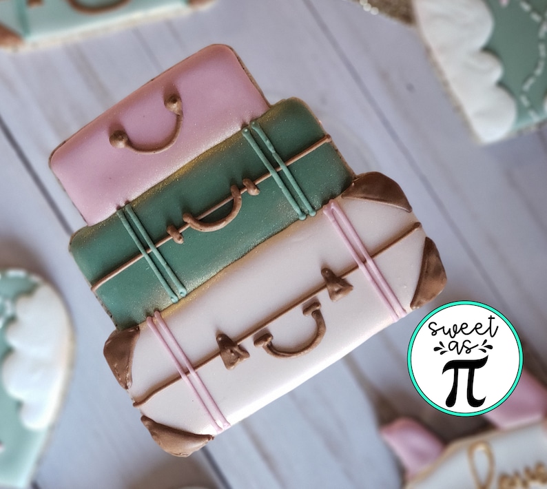 May include: A stack of three decorated cookies shaped like suitcases. The top suitcase is pink, the middle suitcase is green, and the bottom suitcase is white. Each suitcase has a brown handle and gold accents.