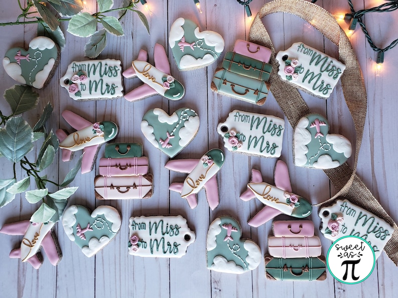 May include: Assortment of decorated sugar cookies with a travel theme. Cookies feature airplanes, luggage, hearts, and clouds. Some cookies have the text "from Miss to Mrs" on them.