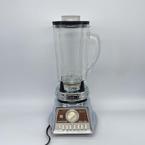 May include: A vintage Waring blender with a clear glass pitcher and a black lid. The base is chrome with a wood-grain panel and control buttons. A power cord extends from the base. The blender is designed for kitchen use.