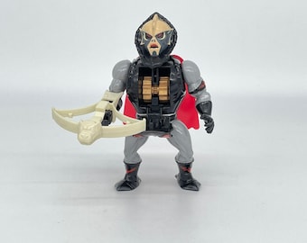 May include: A black and gray action figure with a red cape and a white crossbow. The figure has a detailed head with a black mask and a gray face. The figure is holding the crossbow in its right hand.