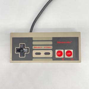 May include: A rectangular beige and dark gray video game controller featuring a black directional pad on the left, two red circular buttons labeled B and A on the right, and two gray buttons labeled SELECT and START in the center with Nintendo text.