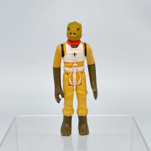 May include: A vintage action figure of a reptilian alien character. The figure is dressed in a yellow jumpsuit with a white vest and a red collar. The figure has brown arms and feet and a reptilian head.