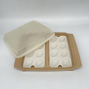 May include: A tan plastic egg tray with a clear lid. The tray has two sections, each with six egg-shaped indentations. The lid is partially open, revealing the white interior of the tray. The tray is designed to hold a dozen eggs.