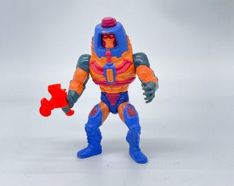 May include: A vintage action figure of a humanoid character with an orange torso, blue legs, and a blue helmet. The figure holds a red weapon. The figure has gray arms and is posed against a white background. A collectible toy.