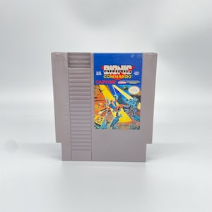May include: A gray Nintendo Entertainment System game cartridge for Bionic Commando. The label features a colorful action illustration of a character with a bionic arm, yellow light beams, and the Capcom logo. The cartridge has a ribbed top edge.