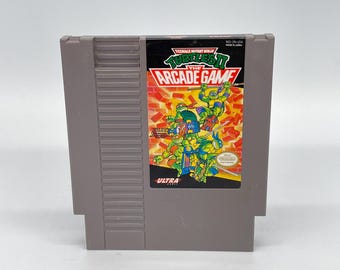 May include: A gray Nintendo Entertainment System (NES) game cartridge for Teenage Mutant Ninja Turtles: The Arcade Game. The label features the game title and images of the turtles. The cartridge is made in Japan.