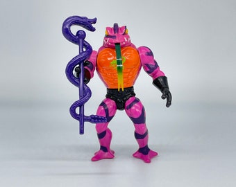 May include: A pink and orange action figure of a frog-like creature with a purple snake staff. The figure has black stripes and a green tongue.
