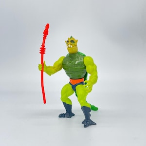 May include: A vintage action figure of a green, muscular humanoid creature with yellow skin, a green cape, and black boots. It holds a red spear. The figure has an orange belt and blue shorts. The toy is posed against a white background.