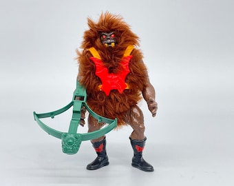 Puede incluir: Una figura de acción de pelo marrón con una pieza de pecho roja y amarilla y una ballesta verde. La figura tiene ojos rojos y una sonrisa de dientes.