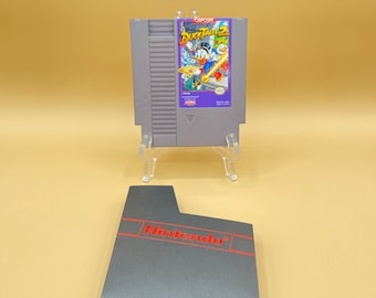 May include: A gray Nintendo Entertainment System (NES) game cartridge for DuckTales 2, featuring a purple and yellow label with a cartoon image of Scrooge McDuck and the text "DuckTales 2". The cartridge is sitting on a clear plastic stand. A gray NES game case with the word "Nintendo" in red is also shown.