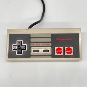 May include: A classic Nintendo controller with a beige and gray color scheme. It features a directional pad, select and start buttons, two red action buttons labeled A and B, and the Nintendo logo. A black cable extends from the top.
