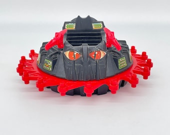 May include: Black and red toy spaceship with red glowing eyes and a black and green cockpit. The spaceship has a red ring around the bottom with black spikes.