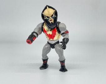 May include: A black and gray action figure with a red and gold chest plate. The figure has a black hooded head with a white face and a red mouth. The figure is holding a black and red weapon in its right hand.