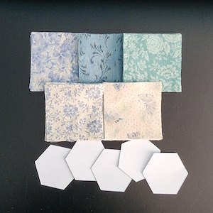May include: Five square fabric swatches featuring various floral and botanical patterns in shades of blue, cream, and teal, arranged in two rows above five plain white hexagonal paper templates used for quilting and fabric crafting projects.